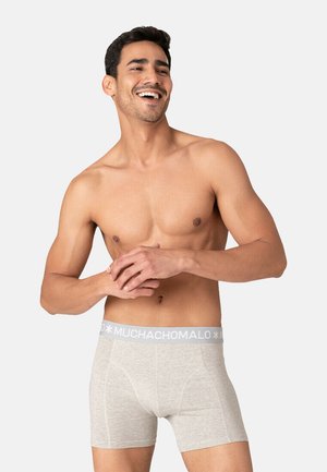 Boxer in cotone grigio con elastico bianco che presenta il testo "MUCHACHOMALO." Modello sorridente, mani incrociate.