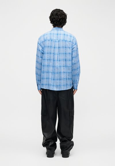 Άτομο με αφρόκριστα μαλλιά φοράει μπλε καρό πουκάμισο με μακριά μανίκια και φαρδιά μαύρη φόρμα cargo, στέκεται με την πλάτη προς τα έξω σε λευκό φόντο.