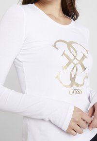 T-shirt bianca a maniche lunghe realizzata in tessuto morbido, con un logo grafico a strisce dorate sul davanti. Scollo rotondo e silhouette aderente.