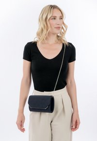Žena v černém tričku s výstřihem do V a béžových kalhotách, nesoucí malou černou kabelku přes rameno se stříbrným řetízkem.