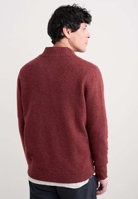 Sarkana teksturēta adīta džemperis ar apkakli, ar ribota tipa aprocēm un apakšmalu, redzams no aizmugures. Valkāts virs gaiša toņa krekla.