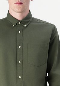 Camicia verde oliva con colletto a punta, tasca frontale e bottoni simili a perle. Realizzata in tessuto morbido con una texture liscia.