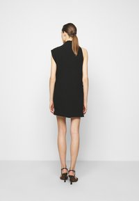 Svart ärmlös klänning med hög krage, skräddarsydd passform och knä-lång nederdel. Bärs med svarta remsandaler. Slät tygtextur.