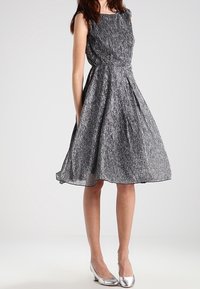 Ärmlös, knälång klänning i strukturerad svart och silver, med en figursydd överdel och utsvängd kjol. Kombinerad med silverfärgade klackar.