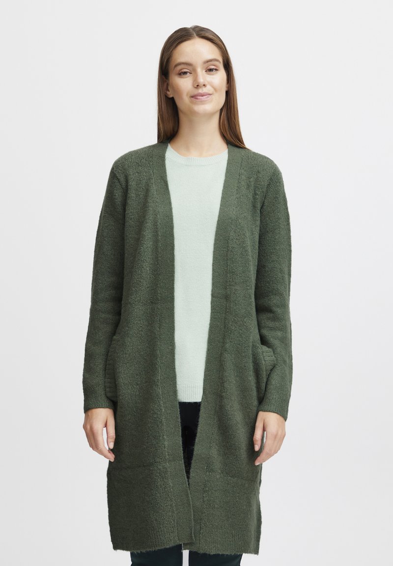 Olive green long cardigan made of textured knit with open front and side pockets, worn over a light mint sweater and dark pants.
