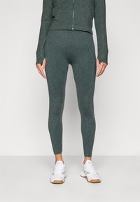 Leggings de sport vert foncé avec une finition texturée et brillante et une coupe ajustée. Associés à une veste zippée assortie et des baskets blanches.