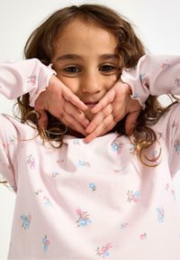 Camicia rosa chiaro a maniche lunghe con motivo floreale in tonalità di blu e rosa, caratterizzata da polsini arricciati. Capelli lunghi e ricci che incorniciano il viso.