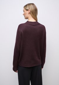 Kvinde, der står med ryggen til, iført en mørk lilla strikket sweater med ribbet krave og manchetter, parret med sorte bukser mod en ensfarvet baggrund.