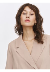 Beige tailored blazer with sharp lapels, structured shoulders, and a smooth texture. Model features soft waves and minimal accessories.