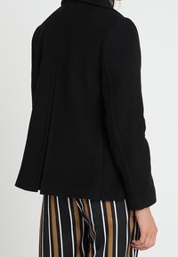 Černý, strukturovaný kabát s vysokým límcem, s křivkovým zadním švem a dlouhými rukávy. Viditelné pruhované kalhoty.