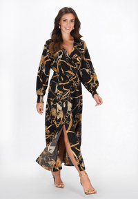 Lang sort kjole med guldkæde og blomsterprint, med lange ærmer, bånd i taljen og et frontslids. Kombineret med gyldne stropper hæle.