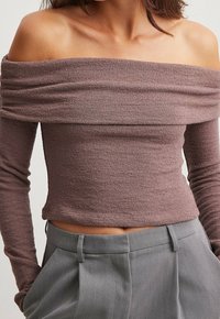 Kvinna som bär en strukturerad, mauve off-shoulder långärmad topp i kombination med högmidjade grå byxor med bältesöglor.