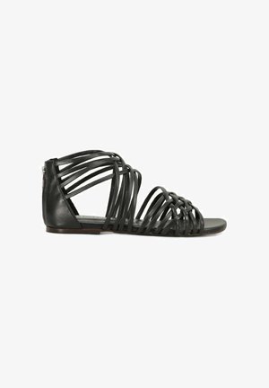 Sandalo nero piatto con molte sottili fasce di pelle e una cerniera sul retro per la chiusura, mostrato da lato su sfondo bianco.