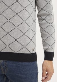 Grå strikket genser med diamantmønster i svart, med en solid svart ribbestrikket kant. Har en liten metalllogo som detalj. Bæres over denim.