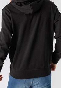 Černá mikina s kapucí vyrobená z měkkého materiálu, s pohodlným střihem, klokaní kapsou a stahovací kapucí. V kombinaci se světle modrými džíny.