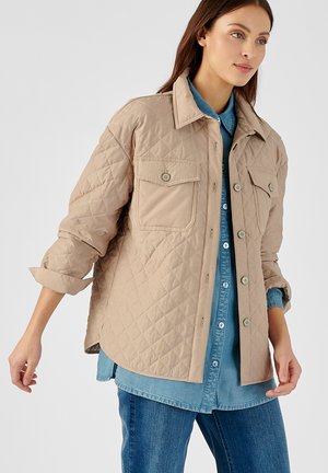 Beige gesteppte Jacke mit Knopfverschluss, zwei Brusttaschen und umklappbarem Kragen, getragen über einem Jeanshemd.
