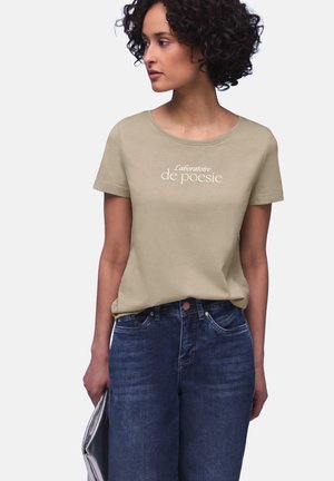 Kvinde med krøllet hår iført en beige "Laboratoire de poésie" T-shirt og blå jeans, som holder en sølvfarvet clutch og ser til siden.