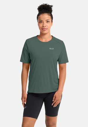 Frau mit lockigem Haar, die ein grünes Kurzarmshirt und schwarze Radsport-Shorts trägt, steht vor einem einfarbigen hellen Hintergrund.