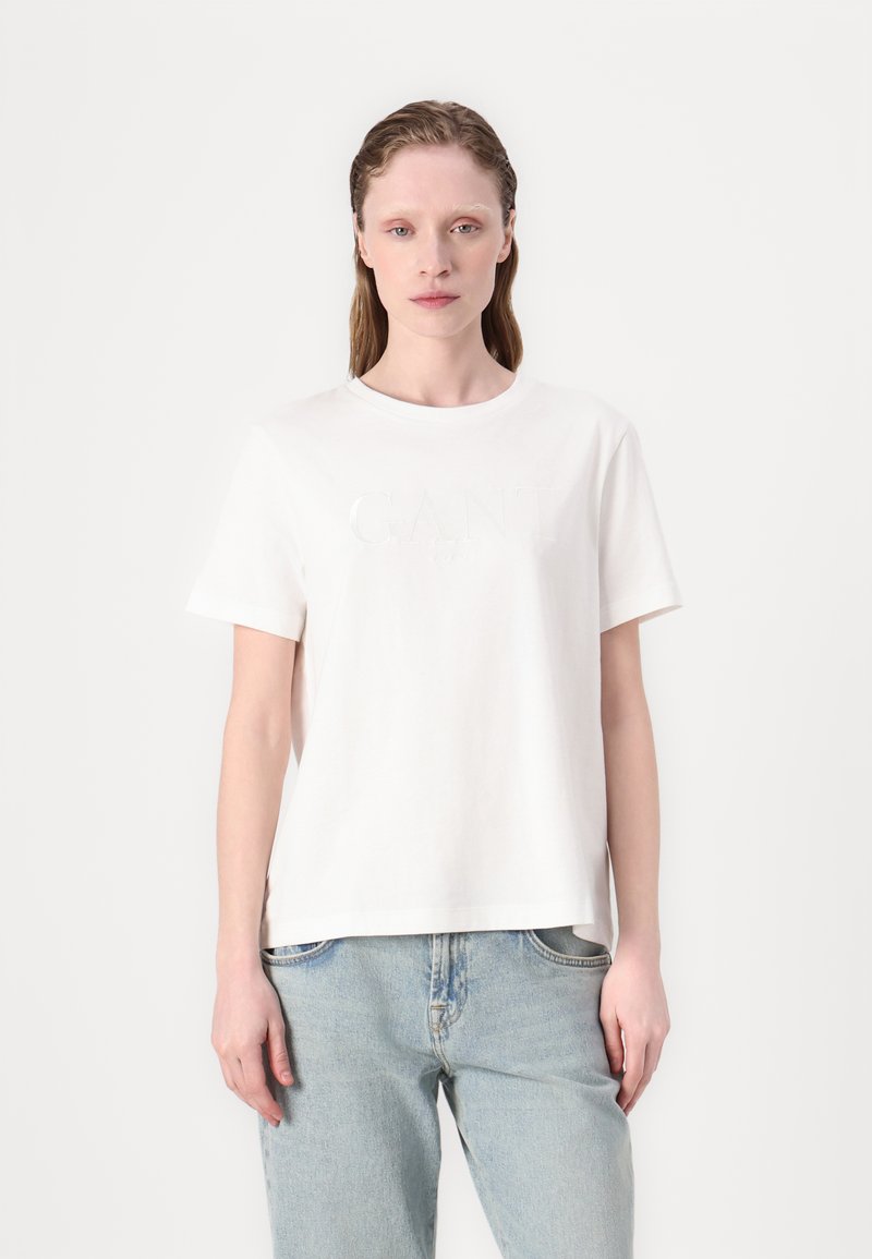 Hvid bomulds t-shirt med korte ærmer, rund halsudskæring og forhøjet GANT-logo på fronten. Kombineret med lyse denim jeans.
