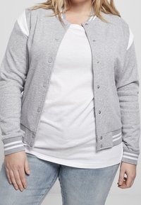Femme portant une veste universitaire gris clair boutonnée par-dessus un t-shirt blanc et un jean bleu, debout devant un fond uni.