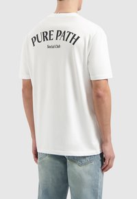 Witte katoenen T-shirt met korte mouwen, met zwarte tekst op de achterkant die "PURE PATH Social Club" in een gebogen ontwerp leest.