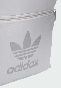 Bijela teksturirana torba s istaknutim sivo-črnimi Adidas logom i dizajnom s tri pruge, s patentnim zatvaračem i glatkim naglascima.