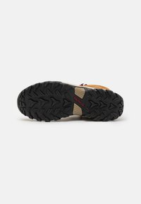 Buty typu trail z trwałą czarną gumową podeszwą z głębokim bieżnikiem, beżową cholewką z perforowanymi detalami i czerwonymi akcentami.