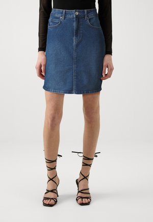 Denim skirt in medium blue with a fitted silhouette, front pockets, button closure, and a hem that reaches mid-thigh, paired with strappy black heels.