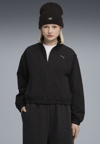 Ung person iført sort Puma-hue, sort halv-zip jakke med Puma-logo, store hoops øreringe og sorte bukser foran en grå baggrund.