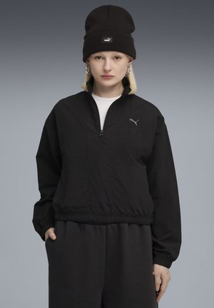 Ung person iført sort Puma-hue, sort halv-zip jakke med Puma-logo, store hoops øreringe og sorte bukser foran en grå baggrund.