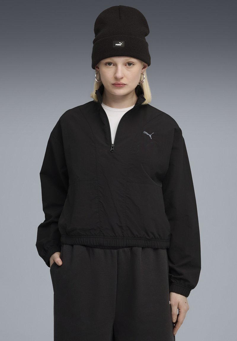 Ung person iført sort Puma-hue, sort halv-zip jakke med Puma-logo, store hoops øreringe og sorte bukser foran en grå baggrund.