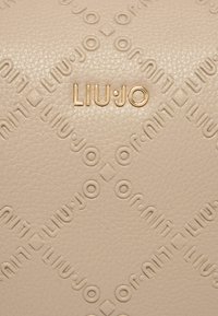 Borsa a tracolla in pelle beige chiaro con motivo del nome del marchio in rilievo e un accento in oro metallizzato sul davanti. Superficie testurizzata.