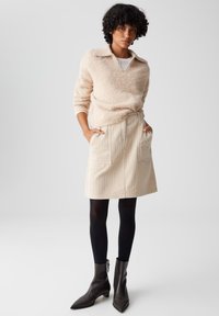 Beige fuzzy sweater met een kraag, gecombineerd met een crèmekleurige geruite rok met voorzakken. Zwarte panty’s en puntige enkellaarzen maken de look af.