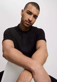 Czarna koszulka polo z zapięciem na guziki, krótkimi rękawami i ściągaczami na mankietach. Model siedzi z założonymi rękami, prezentując krój koszulki.