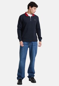 Maglione a coste blu navy con colletto rosso, dotato di mezzo zip. Abbinato a jeans azzurri e scarpe nere, posato davanti a uno sfondo bianco.