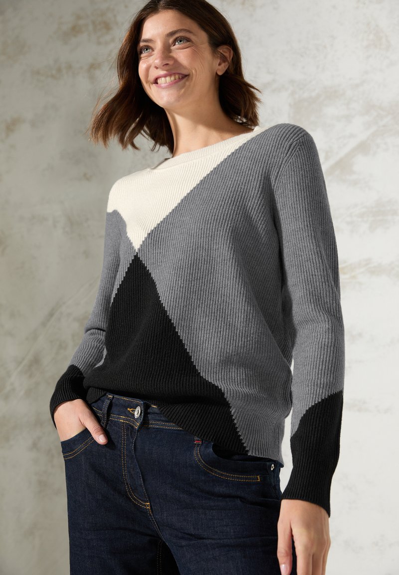 Gestrickter Pullover in Grau, Schwarz und Creme mit einem diagonalen geometrischen Muster, gerippter Textur und langen Ärmeln; kombiniert mit dunklen Jeans.