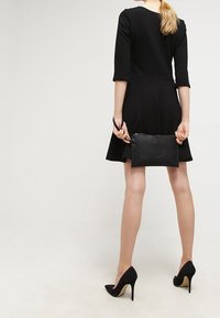 Nainen, jolla on päällä musta mekko ja korkokengät, pitää mustaa clutch-laukkua selän takana, seisten vaalealla taustalla.