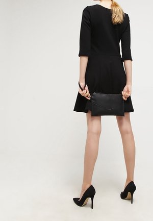 Donna che indossa un abito nero e tacchi alti, tiene una borsa a clutch nera dietro la schiena, in piedi su uno sfondo chiaro uniforme.