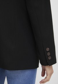 Črn blazer z gladko teksturo, ki vsebuje detajl z dvema gumboma na manšeti. Prilagojen kroj s čistimi linijami. Nosen čez modre kavbojke.