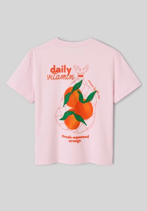 T-shirt rose avec graphique au dos de trois oranges dans un filet, feuilles vertes, et texte orange : « vitamine quotidienne », « pressé par le soleil », « orange fraîchement pressée ».