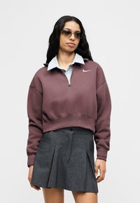 Μπορντώ κοντή φούτερ με φερμουάρ στον γιακά και λογότυπο Nike, συνδυασμένη με γκρι πλισέ φούστα. Φορεμένη πάνω από ανοιχτό μπλε πουκάμισο με γιακά.