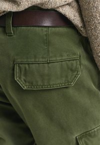 Grüne Stoffhosen mit einer rechteckigen Gesäßtasche und Stichdetails. Das Label "GANT" ist dezent über der Tasche eingprägt.