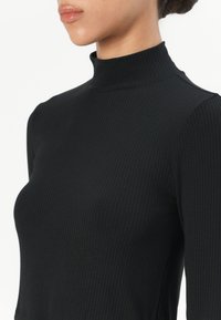 Svart ribbet høyhalset topp med en tettsittende design, høy krage og lange ermer. Laget av et mykt, elastisk stoff for komfort.