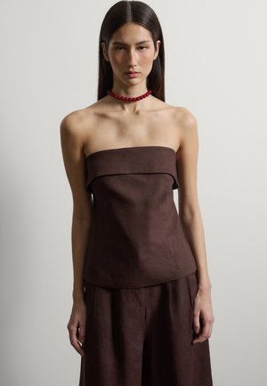 Junge Frau mit langen dunklen Haaren, die ein schulterfreies, dunkelbraunes Oberteil und eine Hose sowie eine auffällige rote Perlenkette trägt, vor einem neutralen Hintergrund.