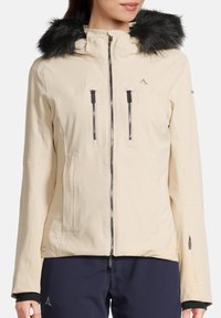 Beige wasserdichte Jacke mit abnehmbarem schwarzen Kunstfellkapuze, ausgestattet mit zwei mit Reißverschluss versehenen Brusttaschen und einem Reißverschluss vorn.