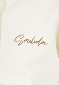 Witte hoodie met een gouden geborduurd "Smilodon" logo. De stof is glad met een zachte textuur en heeft een voorzakje.