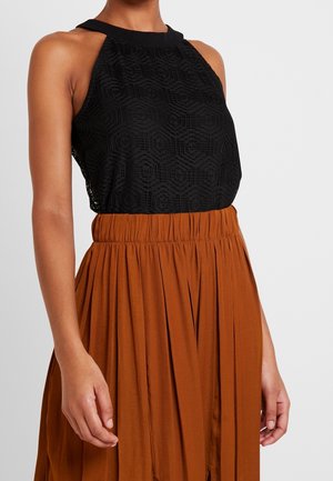 Woman wearing a black sleeveless lace top with high neckline tucked into a rust-colored pleated skirt with gathered waistband.
