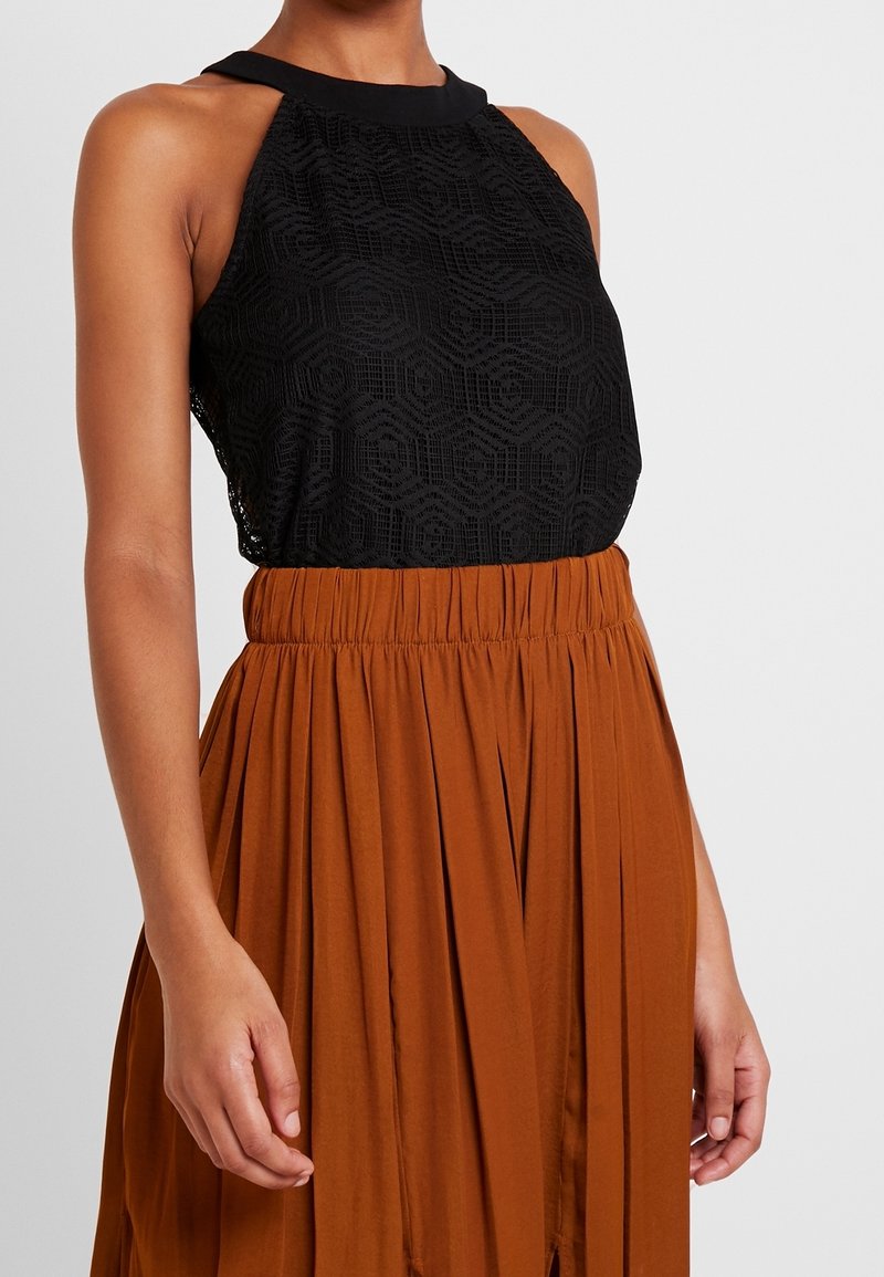 Femme portant un haut en dentelle noir sans manches avec un col montant, rentré dans une jupe plissée couleur rouille avec une taille froncée.
