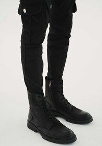 Černé vojenské boty z kůže s designem na tkaničky, bočním zipem a odolnou podrážkou. Nošeny s černými přiléhavými kalhotami.