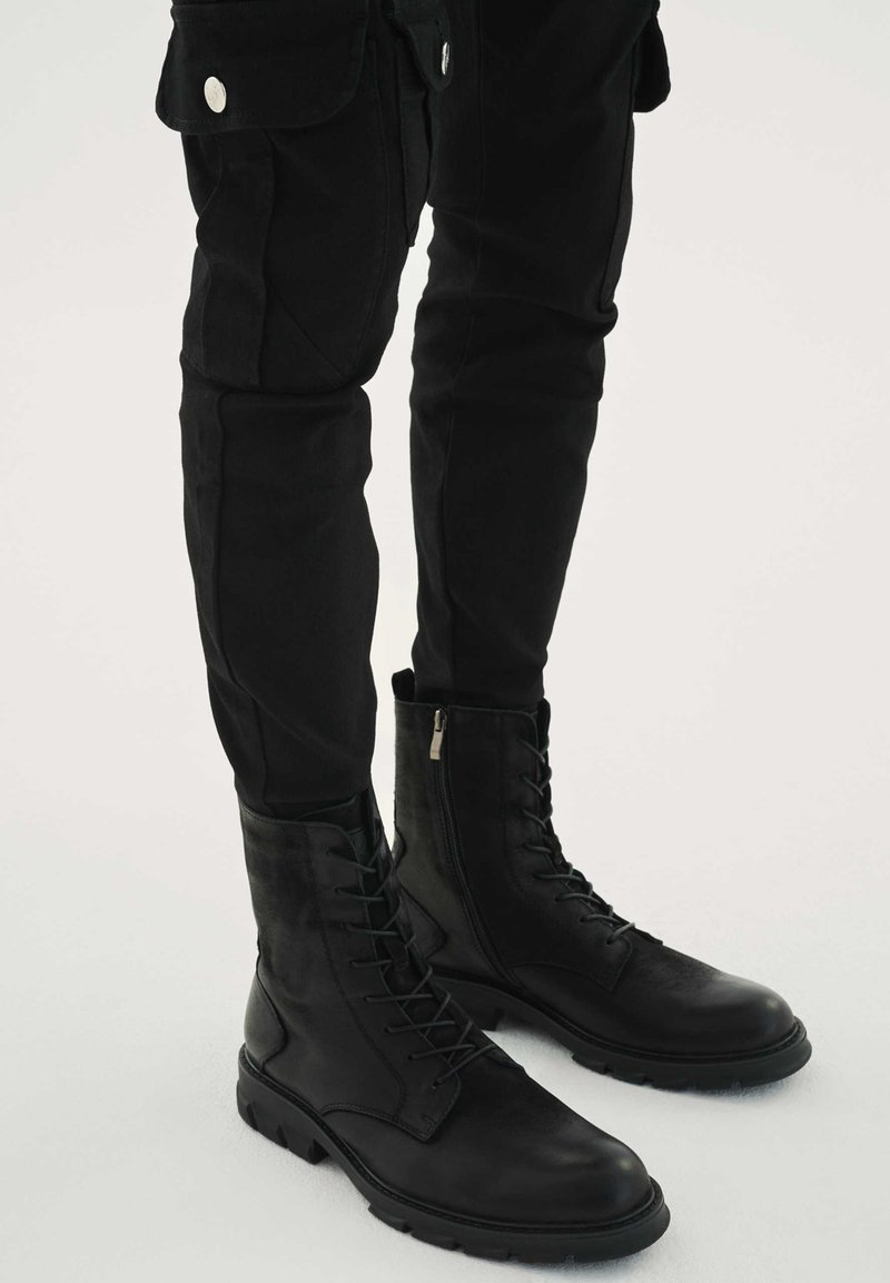 Černé vojenské boty z kůže s designem na tkaničky, bočním zipem a odolnou podrážkou. Nošeny s černými přiléhavými kalhotami.